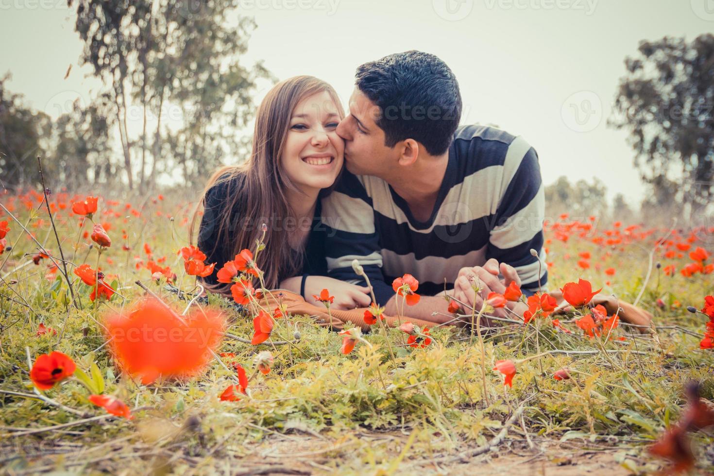 Man kissing woman and she has a toothy smile while they laying on the grass in a field of red poppies photo