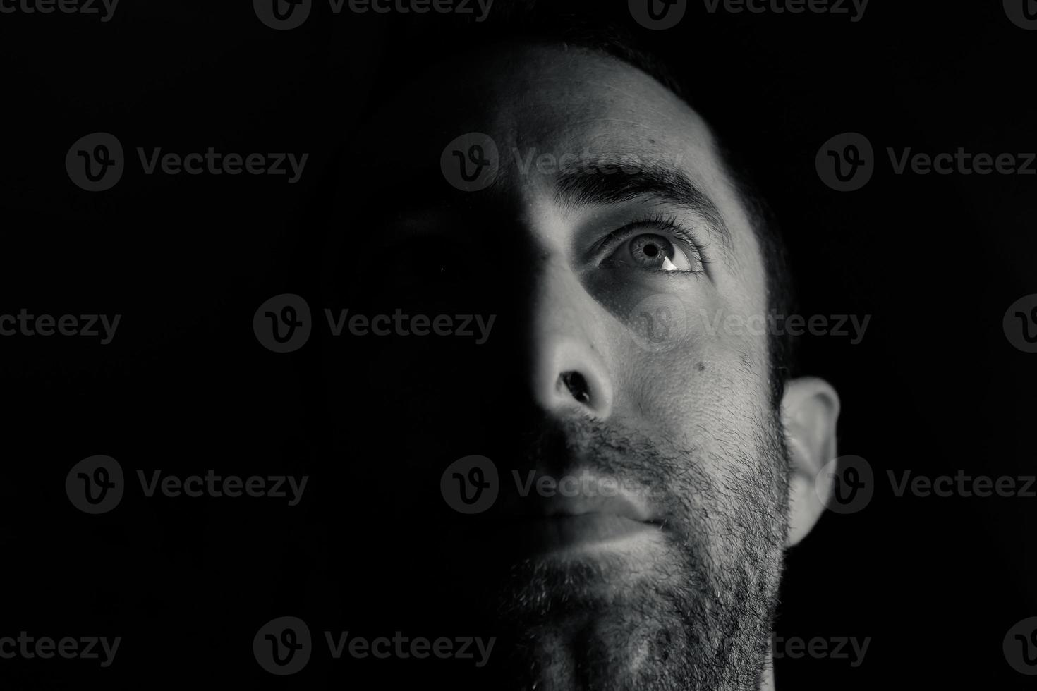 Dramático retrato de un joven medio rostro inexpresivo en la sombra profunda mirando hacia arriba aislado sobre fondo negro foto