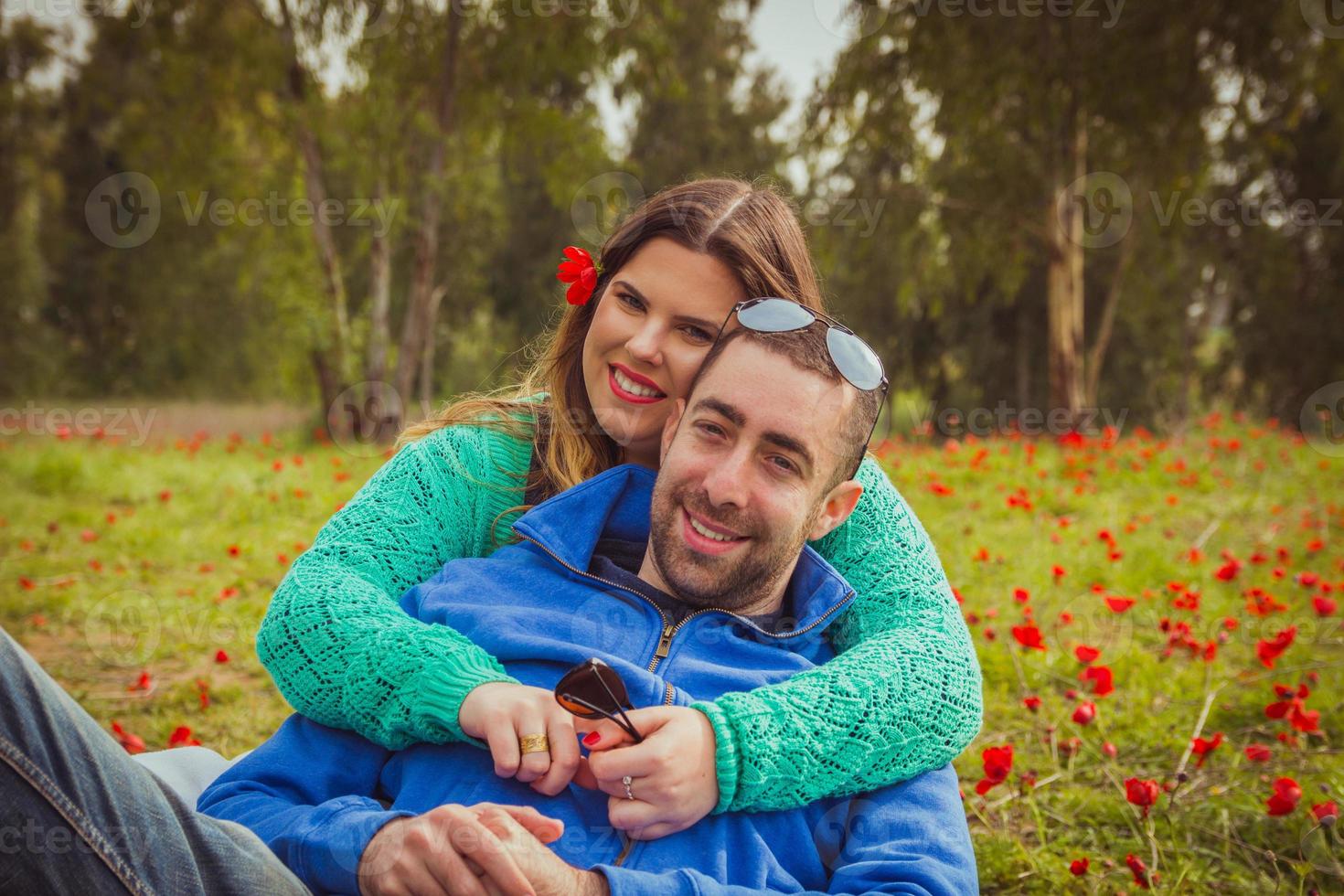 Pareja joven sentada en la hierba en un campo de amapolas rojas y sonriendo a la cámara foto