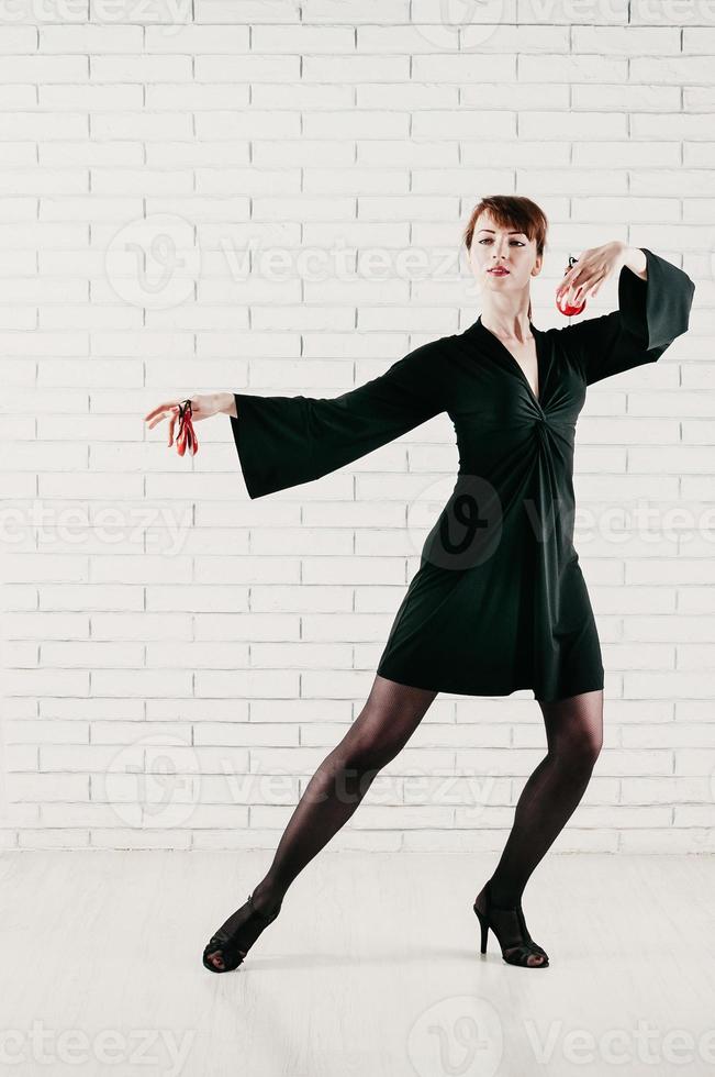 Atractiva mujer joven en vestido negro, bailando con castañuelas rojas, fondo de ladrillo blanco. foto