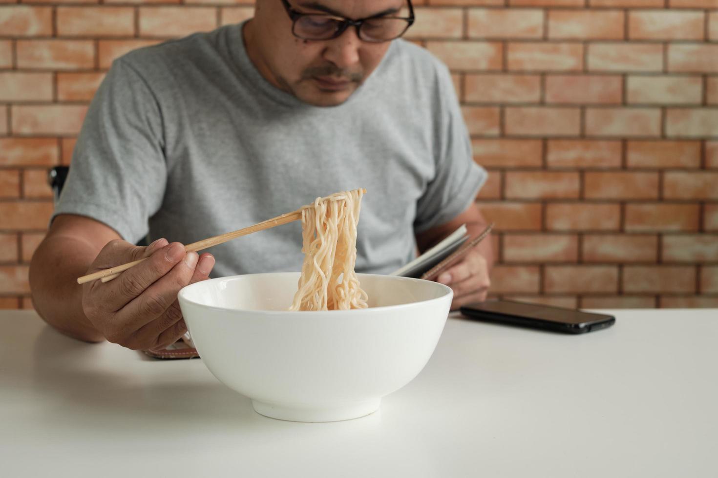 Asian male worker reads an appointment book while eating instant noodles in white bowl with chopsticks on table in brick wall background office during a lunchtime break, a hastily unhealthy lifestyle. photo