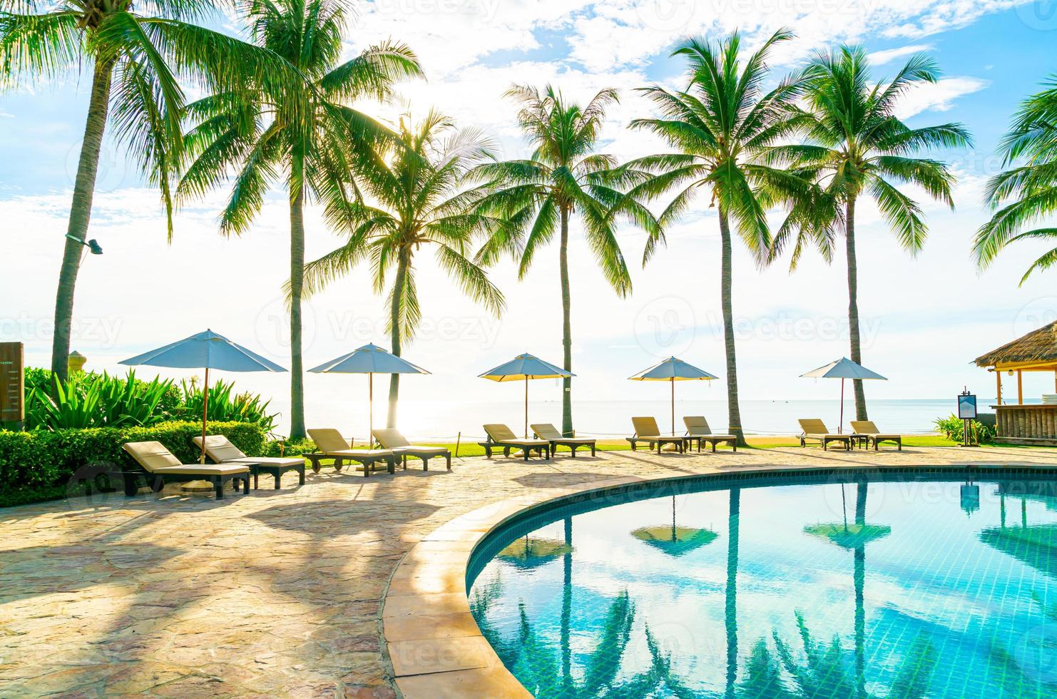 Hermosa sombrilla de lujo y una silla alrededor de la piscina al aire libre en el hotel y resort con palmera de coco en el cielo azul foto