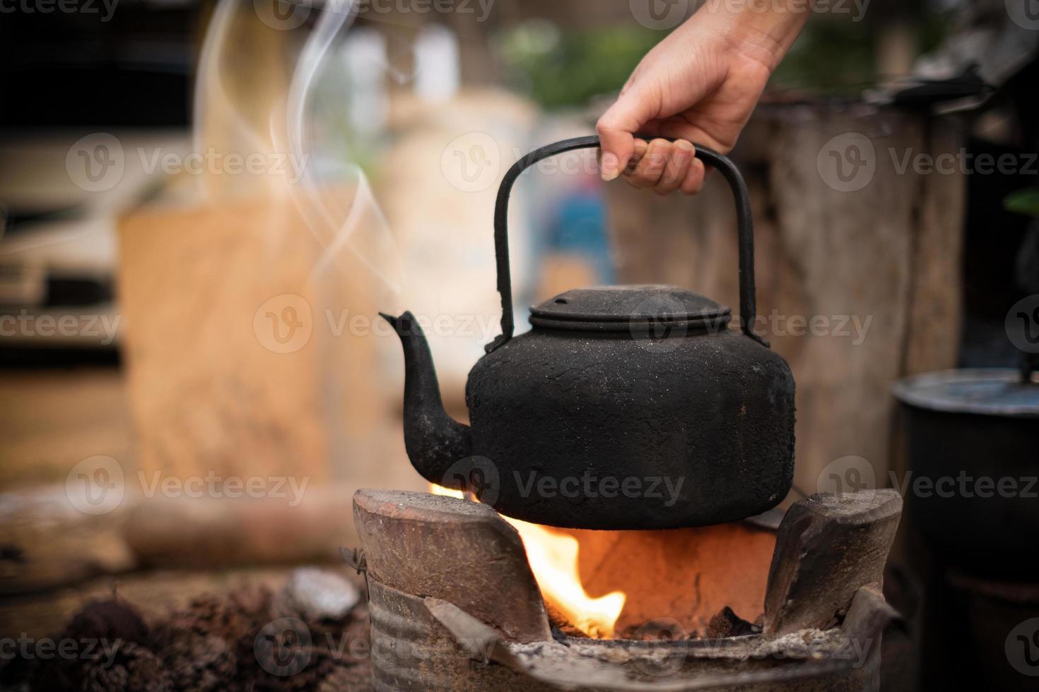 Cerrar mano sujetando hervir agua vieja tetera en el fuego con una estufa de carbón en el fondo borroso foto