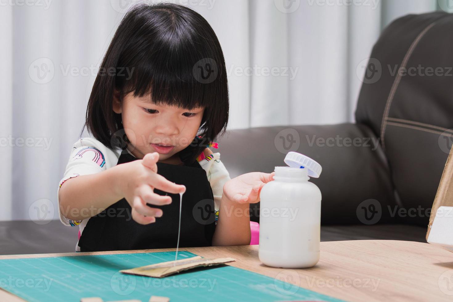 Inventor girl is thrilled to apply stretchy glue onto cardboard pieces out of cardboard box. Child have fun with creative design work pieces. Kindergarten wearing black apron is studying in classroom. photo