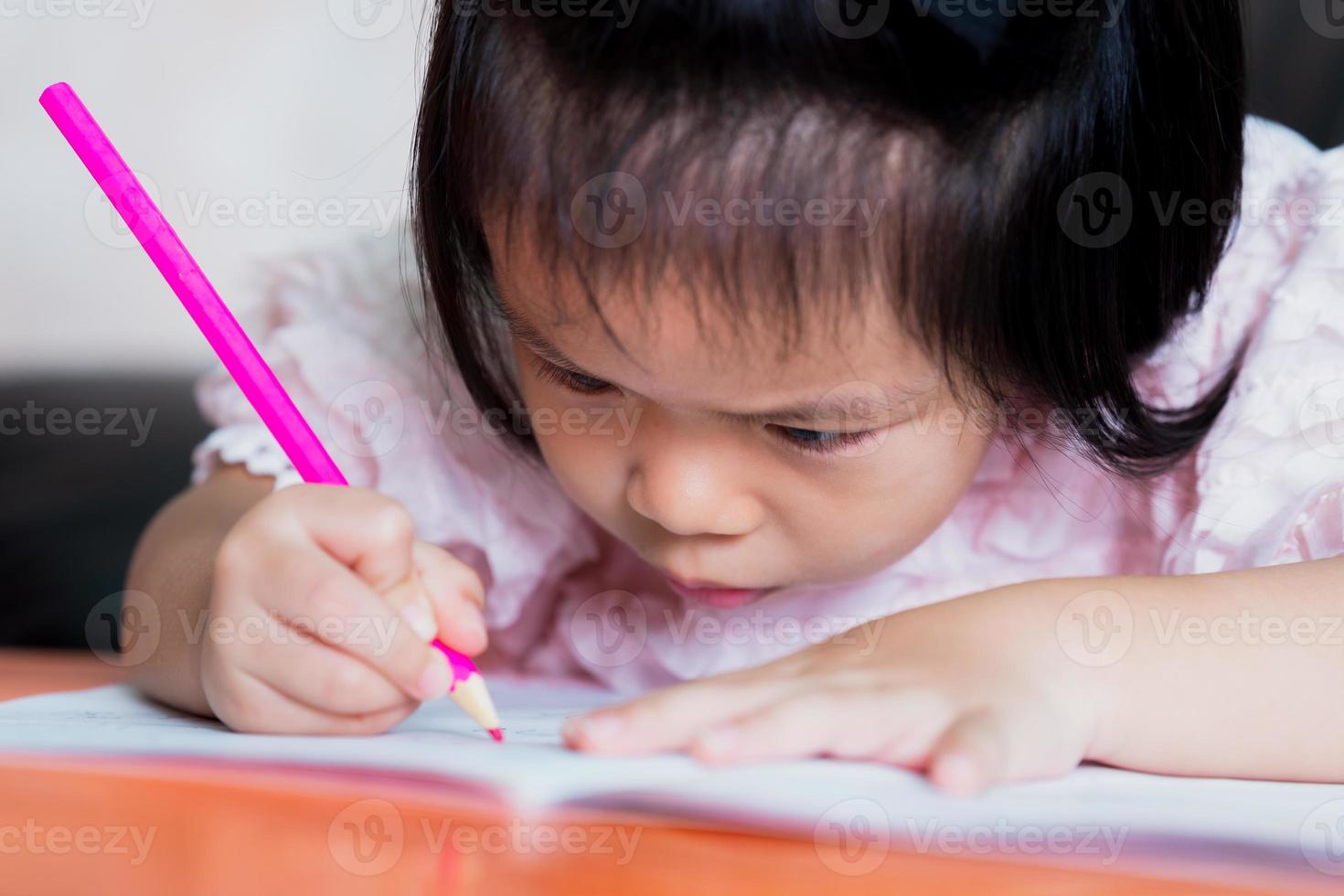 linda chica asiática está coloreando una madera de color rosa en un libro. niño miró de cerca. concepto de postura sentada y problema con los ojos. foto