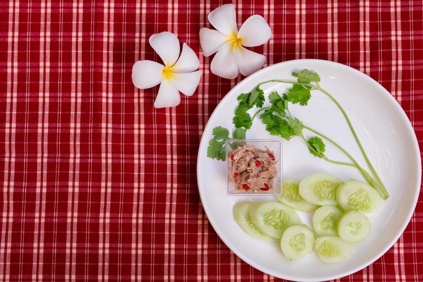 La ensalada de atún yum es pequeña en un plato blanco, adornada con melones y cilantro verde fresco. Fondo de tela escocesa roja y blanca. estilo vintage. decorado con flores de frangipani. foto