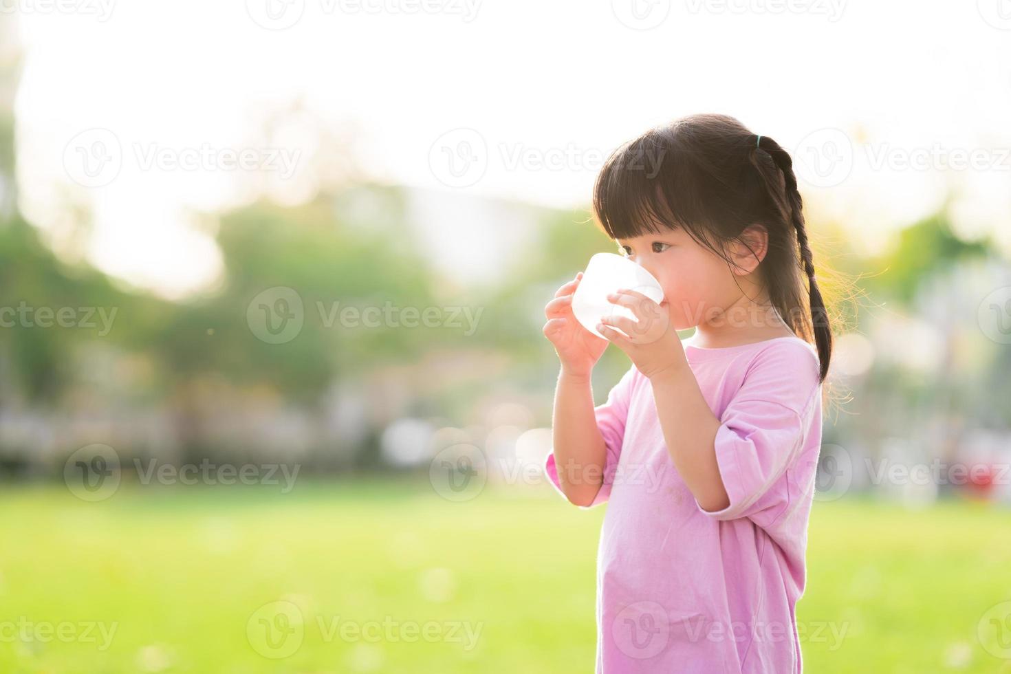 Adorable Asian child girl are drinking fresh water from plastic glasses. Green natural background. In the summer or spring. Side view of kid is 4 year old wearing pink shirt thirsty. photo