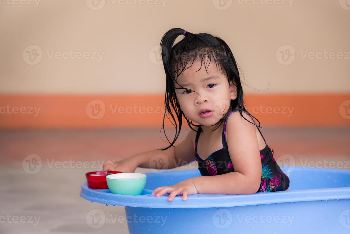 Little Asian girl in bathing suit is playing in blue basin. Play with small glasses. Kid look at camera. Toddler play in water in front yard of country house. Cute child play in water and have fun. photo