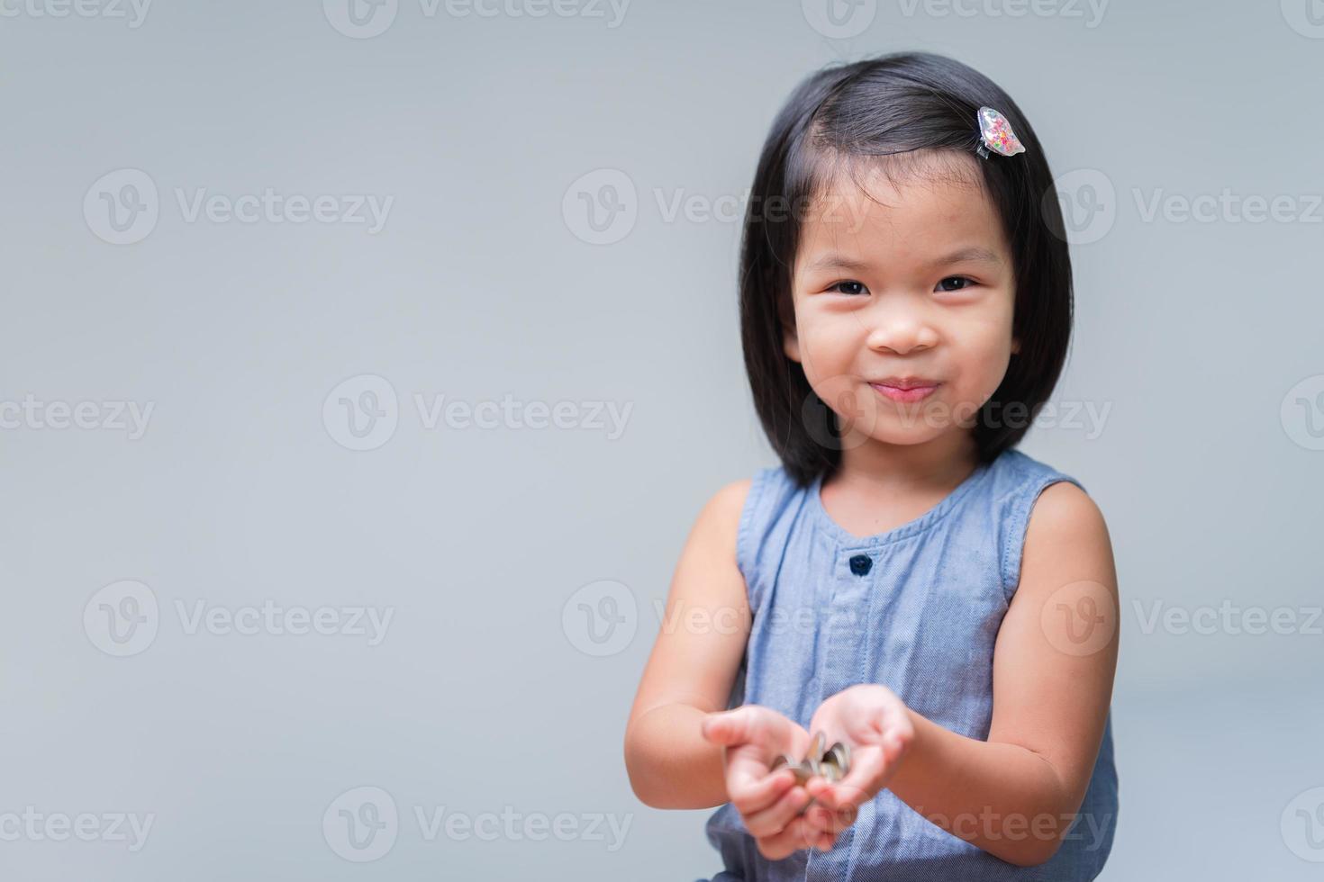 Cute Asian child girl holding coins in her hands. Sweet smiling kid. Happy children playing silver coins. Baby looking at camera. Childhood savings concept. Clean background. Copy space. photo