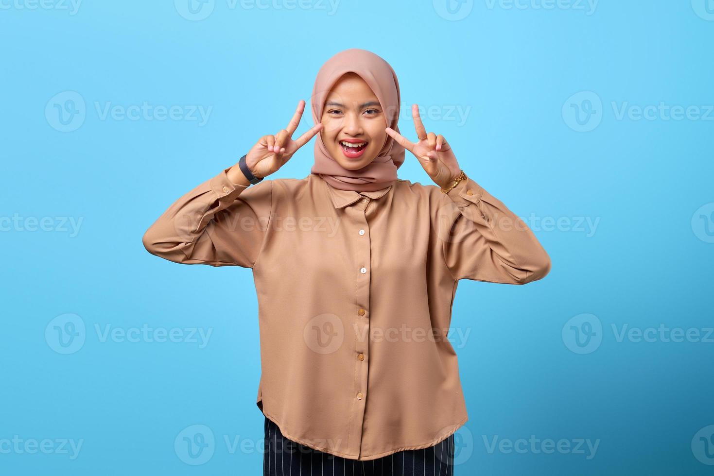Retrato de joven alegre mujer asiática haciendo el signo de la paz con las manos del dedo sobre fondo azul. foto