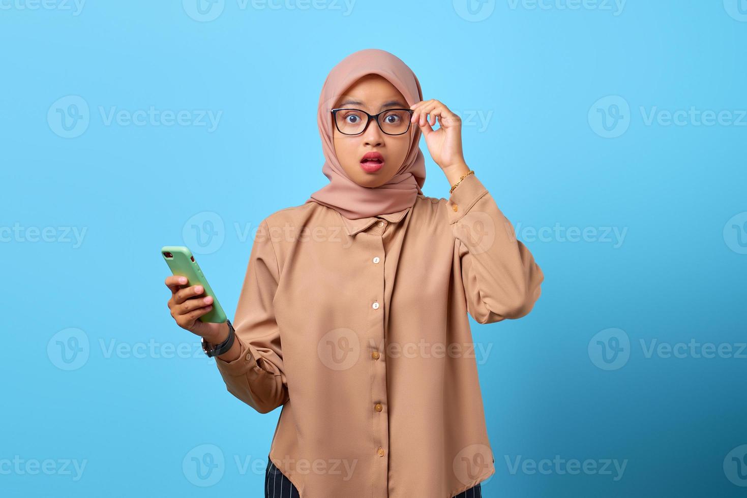 Portrait of surprised young Asian woman holding mobile phone and touching glasses with finger photo