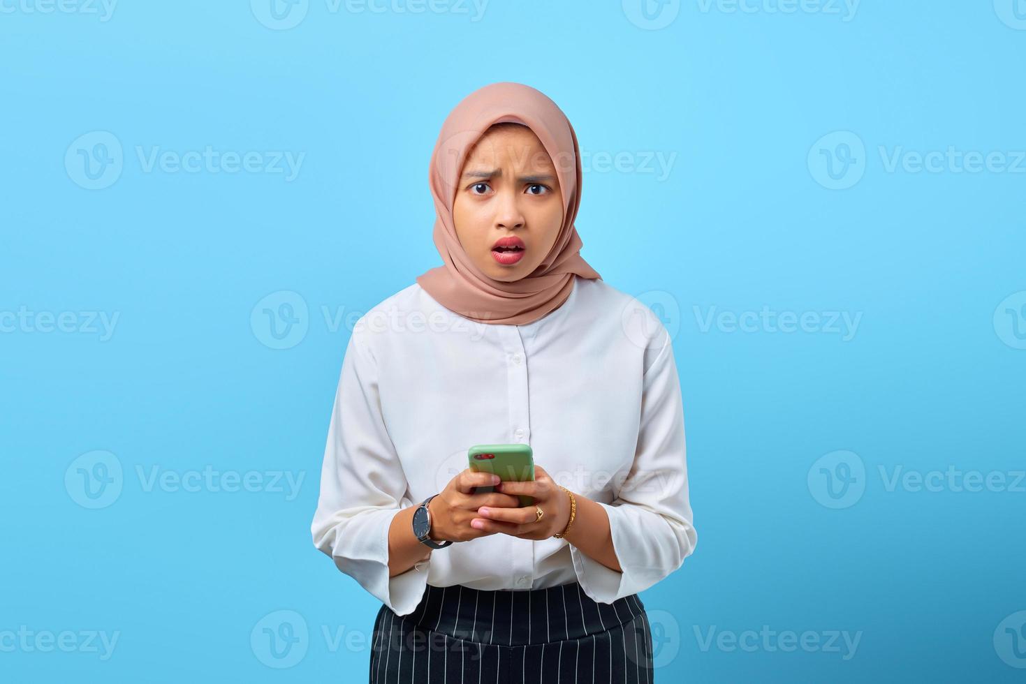 Retrato de mujer asiática joven conmocionada con la boca abierta mediante teléfono móvil sobre fondo azul. foto