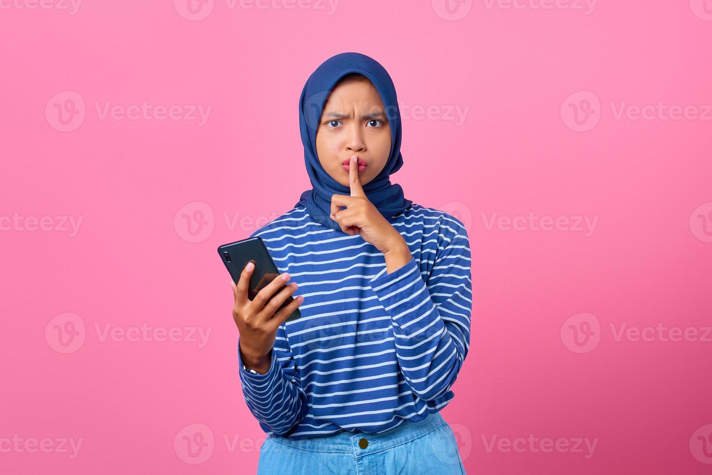 Portrait of young Asian woman holding mobile phone and makes silence gesture with fingers photo