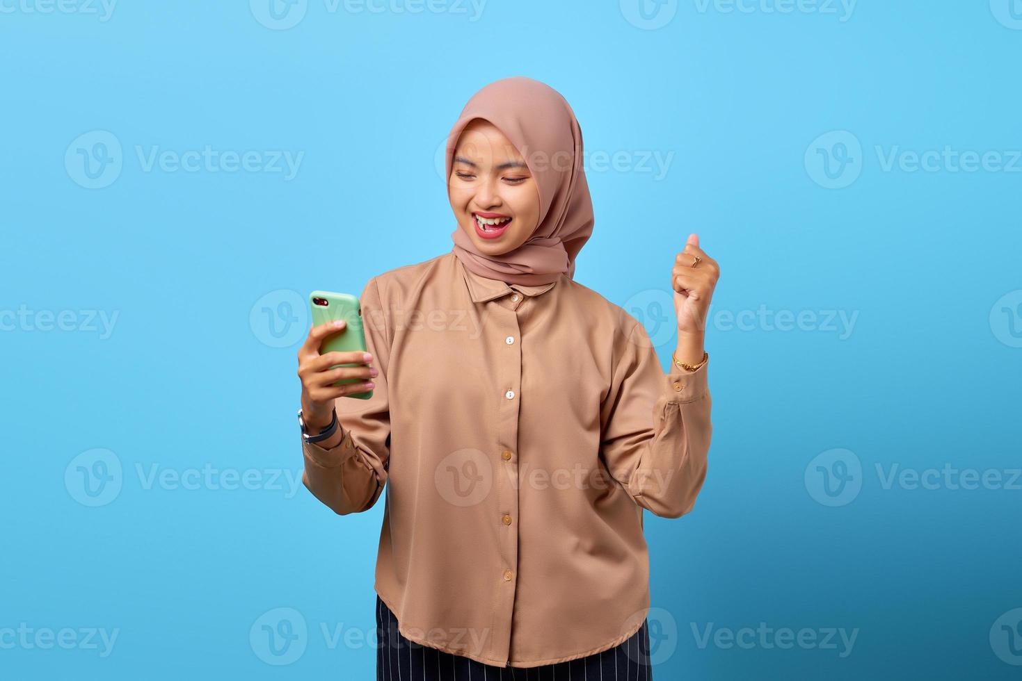 Portrait of excited cheerful young Asian woman celebrating winning with mobile phone on hand photo