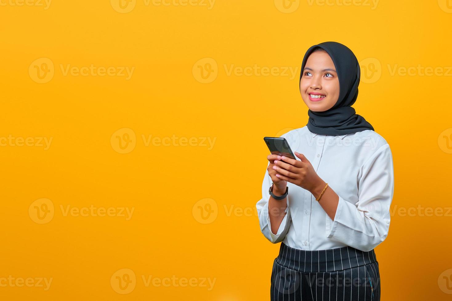Sonriente joven mujer asiática con un teléfono móvil y mirando hacia los lados sobre fondo amarillo foto