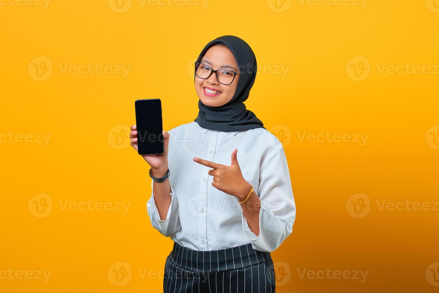 Feliz joven mujer asiática apuntando a la pantalla en blanco del teléfono móvil aislado sobre fondo amarillo foto