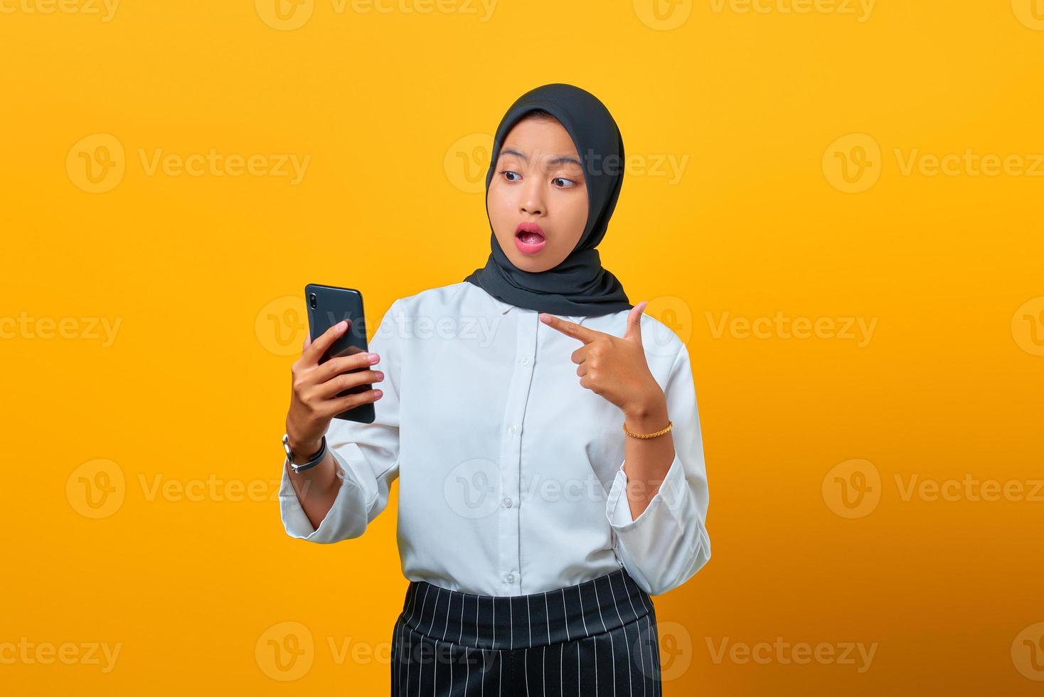 Sorprendido joven asiática apuntando al teléfono móvil aislado sobre fondo amarillo foto