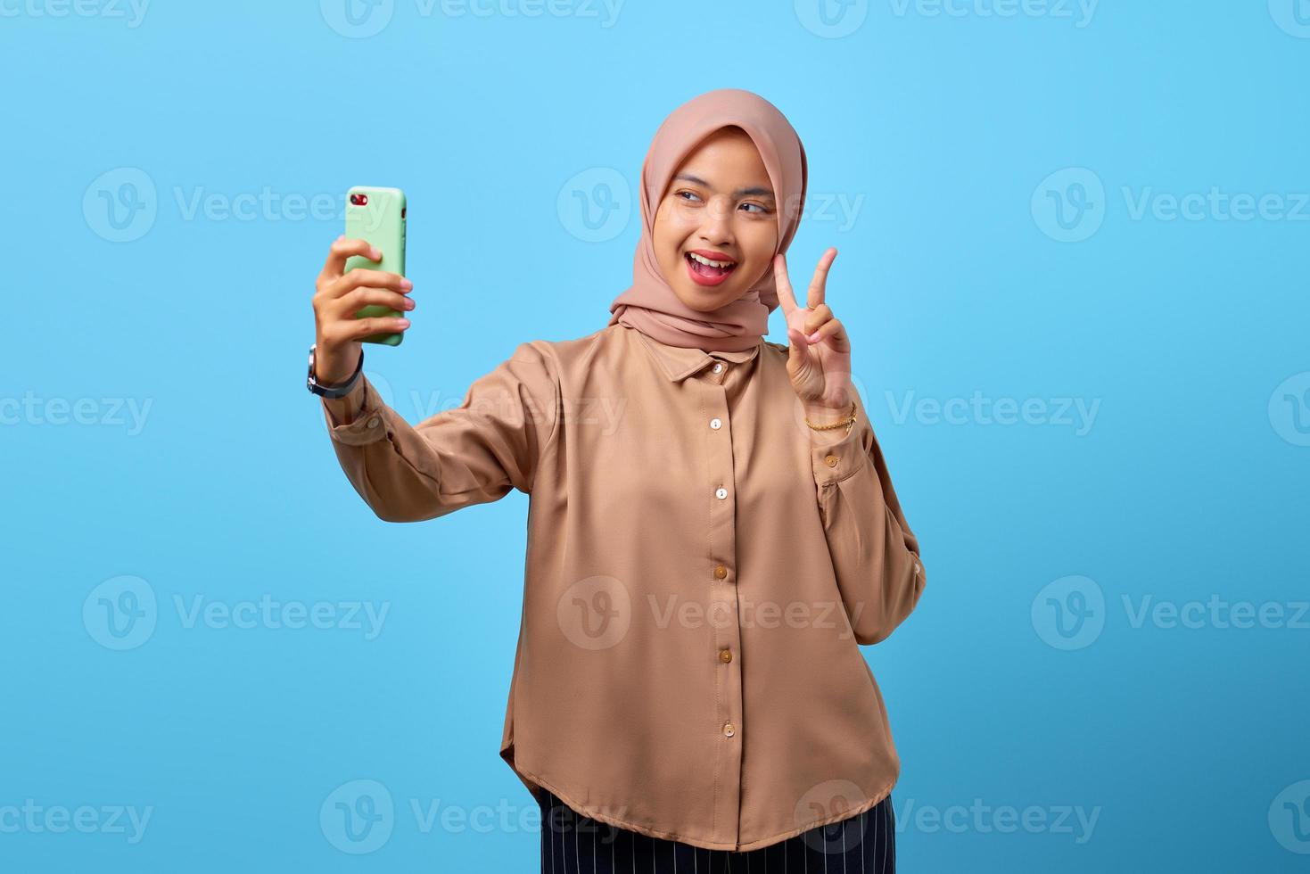 Portrait of cheerful young Asian woman using mobile phone take selfie make peace sign photo