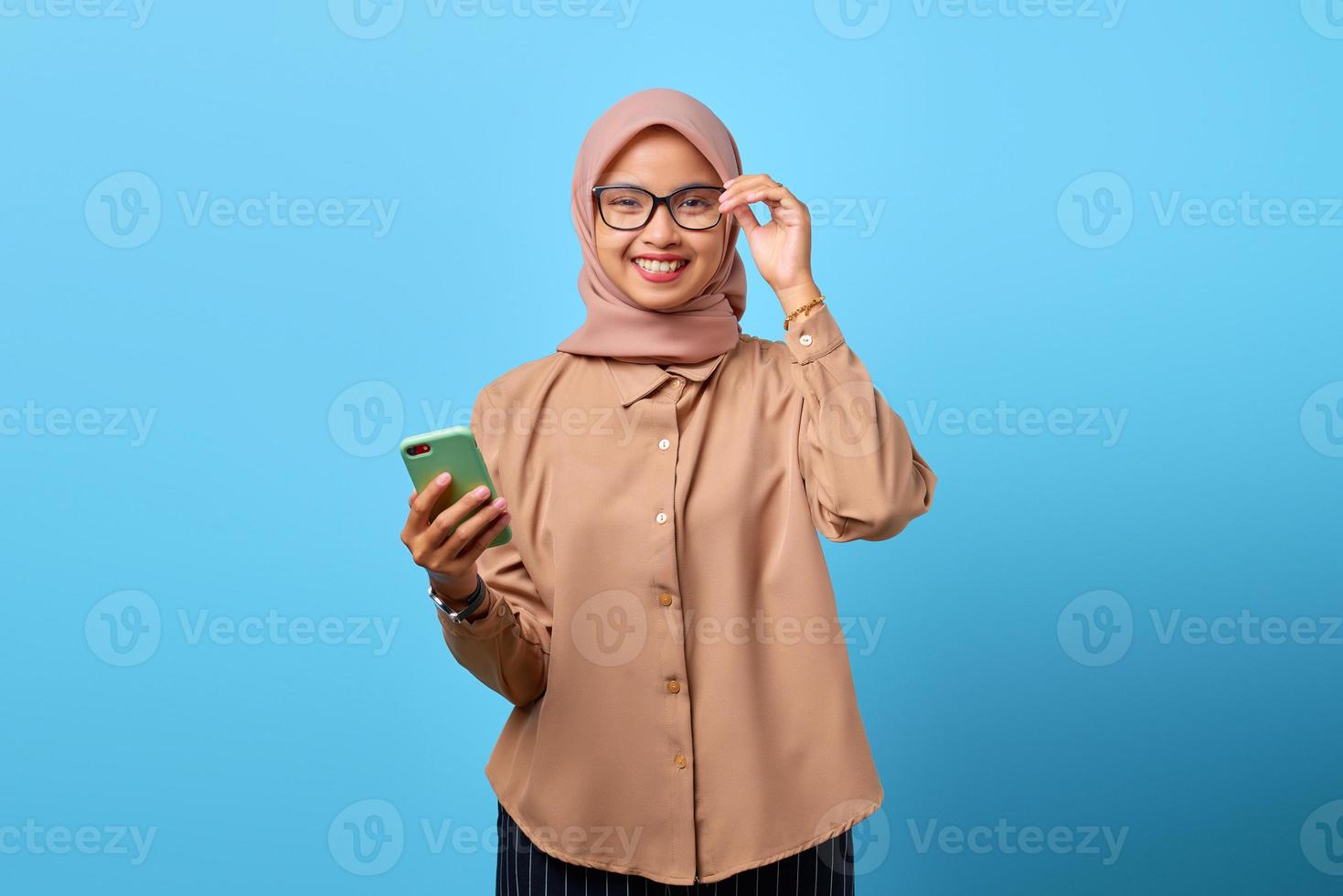 Portrait of smiling young Asian woman holding smartphone and eyewear looking at camera photo