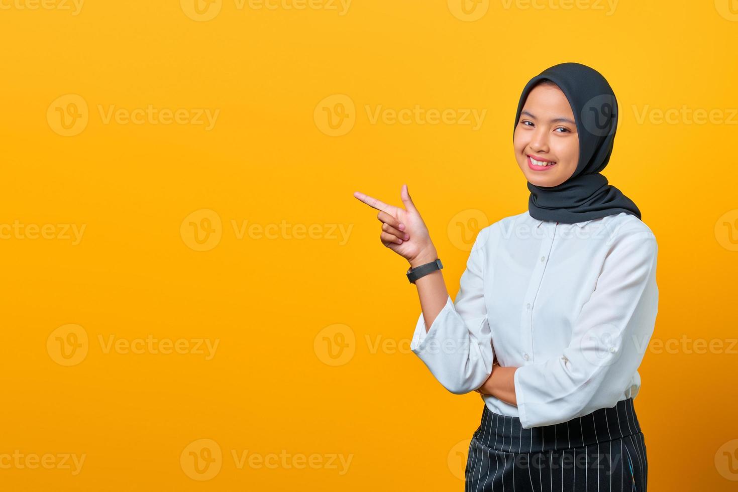 Smiling young Asian woman pointing fingers at copy space on yellow background photo