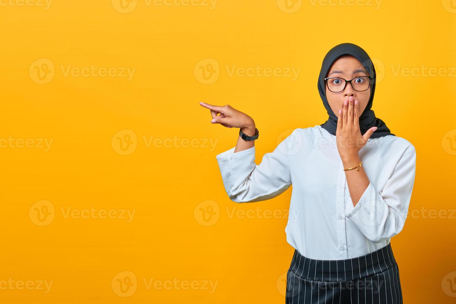 Surprised young Asian woman pointing fingers at copy space on yellow background photo