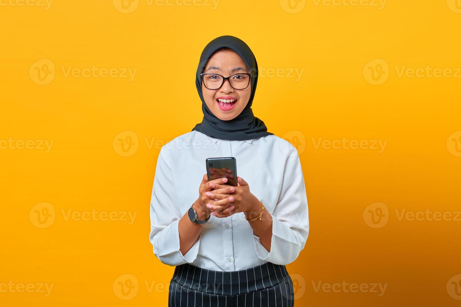 Alegre joven asiática sosteniendo un teléfono móvil y mirando la cámara sobre fondo amarillo foto