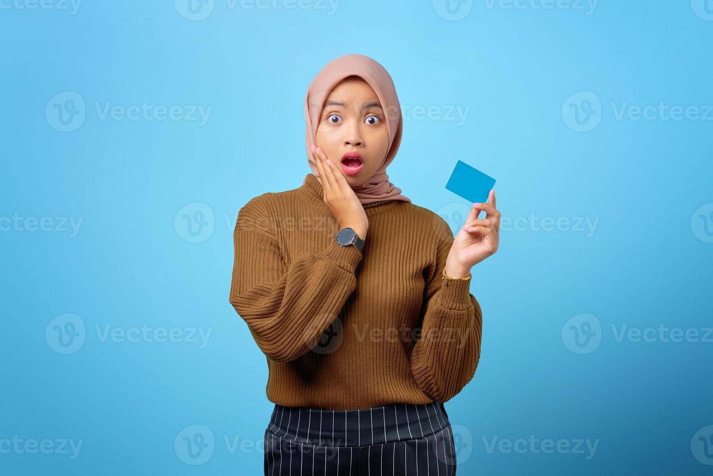 Surprised young Asian woman holding credit card and touch cheeks on blue background photo