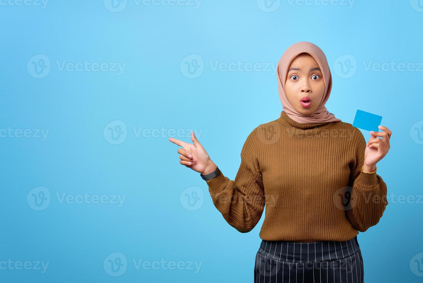 Surprised young Asian woman holding credit card and pointing finger to copy space on blue background photo