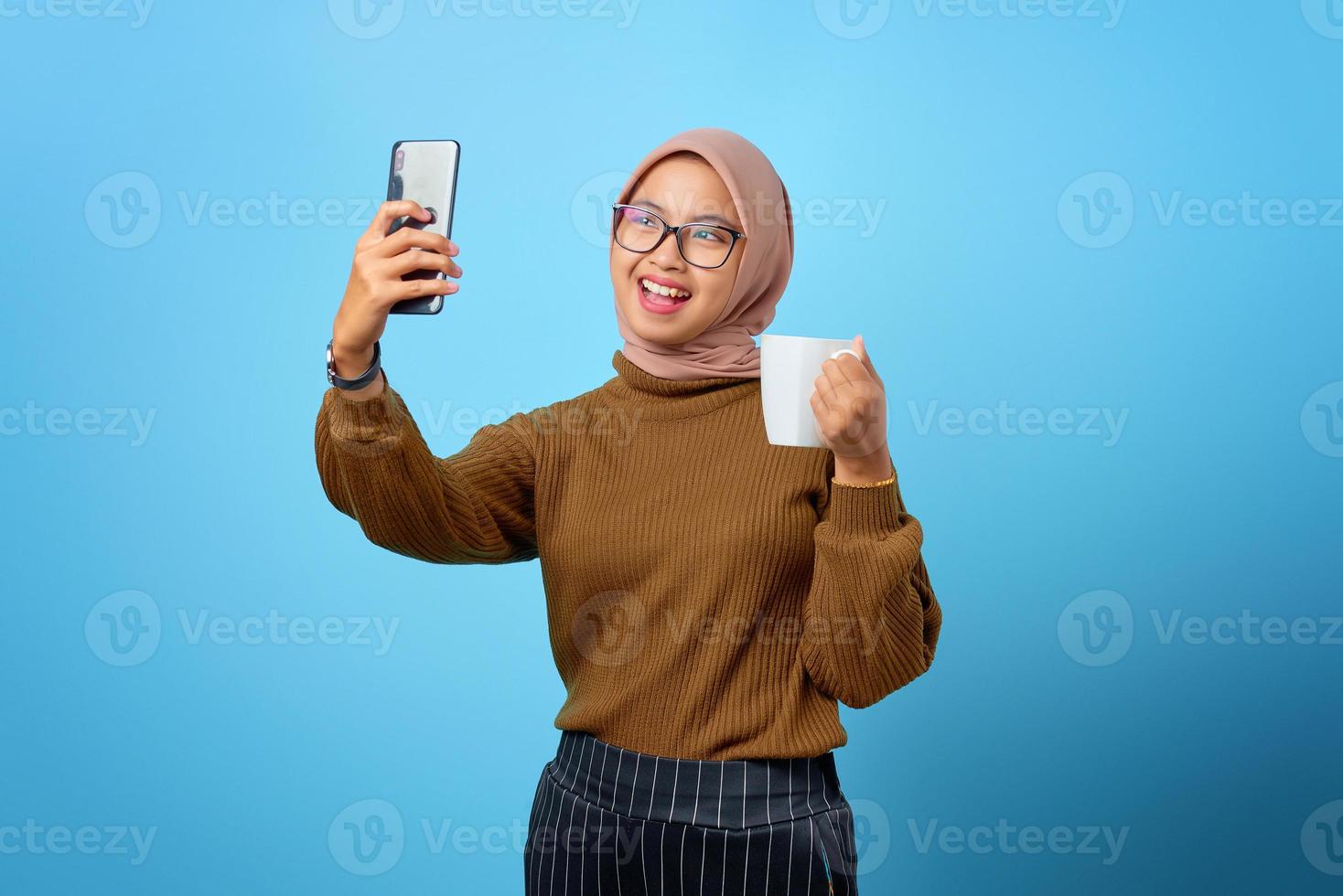 Alegre mujer asiática con teléfono móvil haciendo selfie y sosteniendo la taza sobre fondo azul. foto