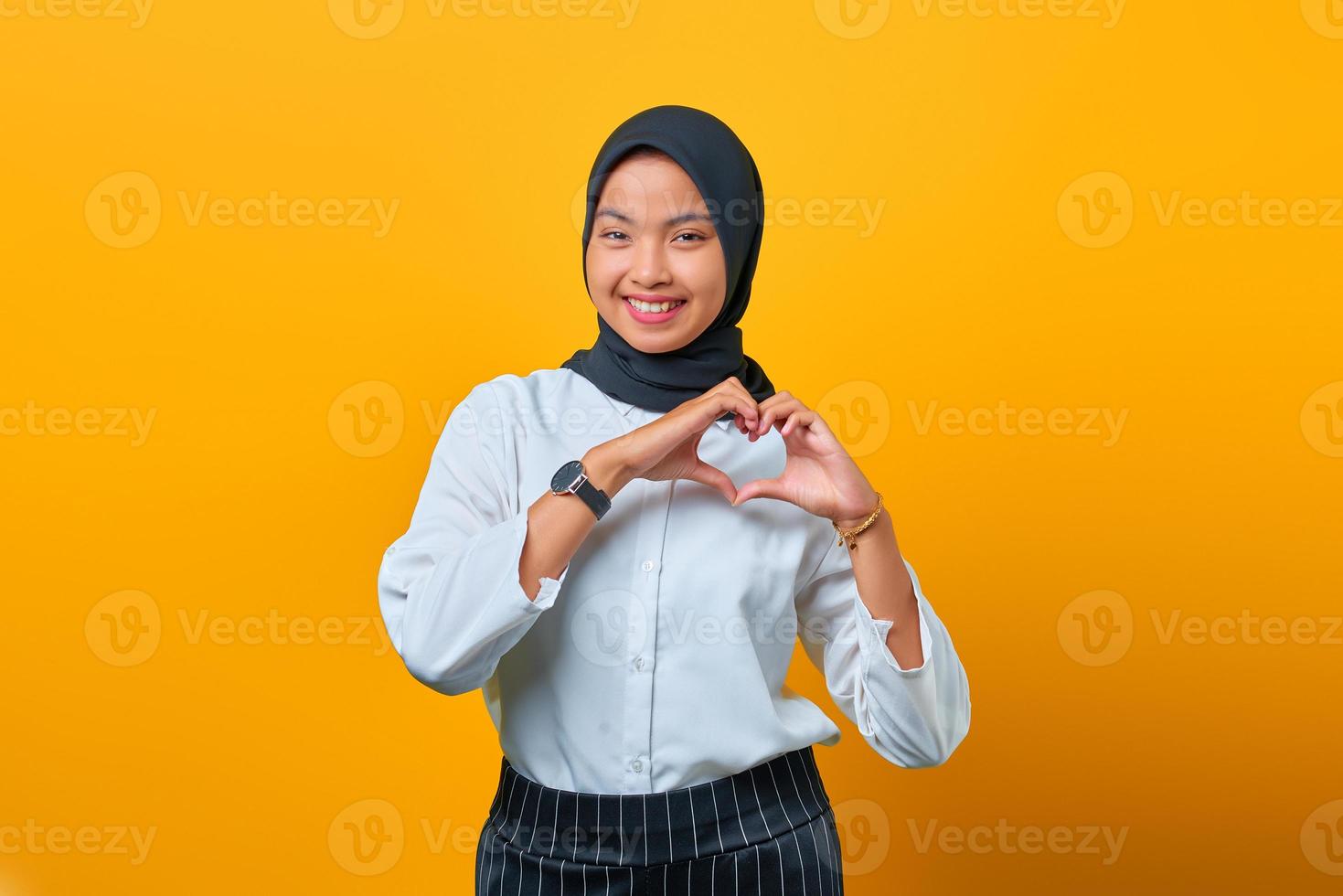 Smiling young Asian woman makes shapes heart sign on yellow background photo