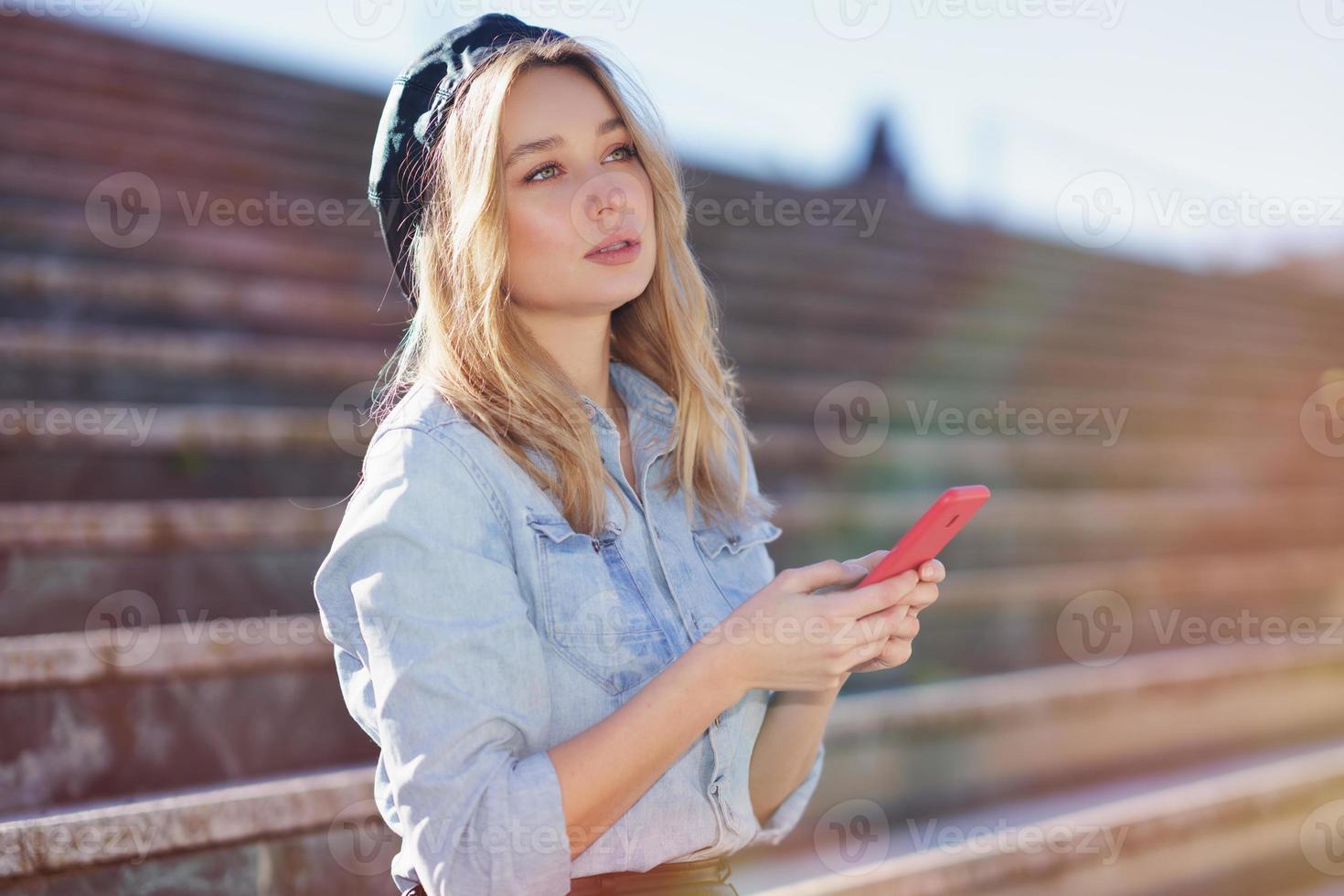 mujer rubia usando un teléfono inteligente sentada en algunos escalones de la ciudad, vestida con una camisa de mezclilla y boina negra. foto