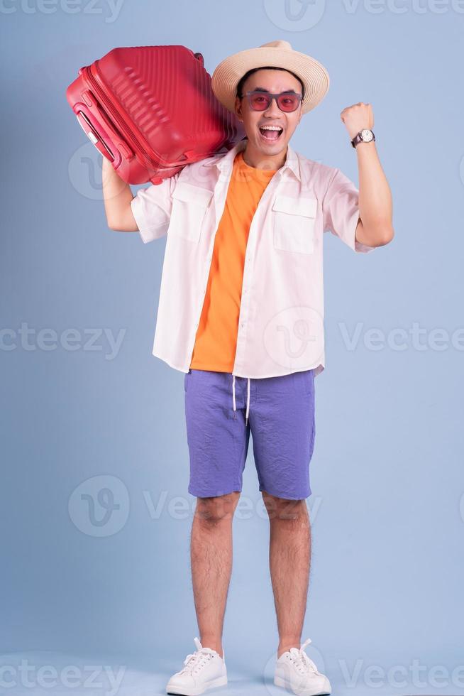 Hombre asiático joven que sostiene la maleta roja sobre fondo azul. foto