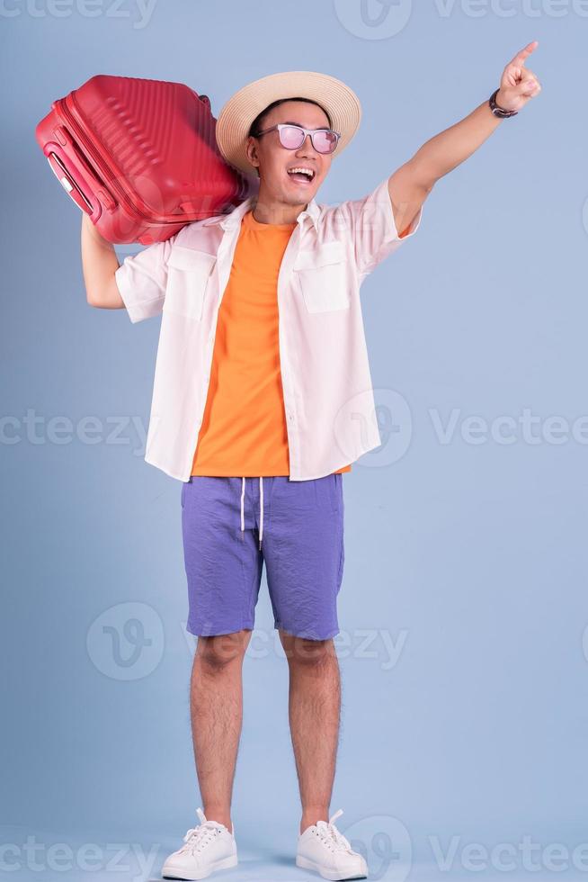 Hombre asiático joven que sostiene la maleta roja sobre fondo azul. foto