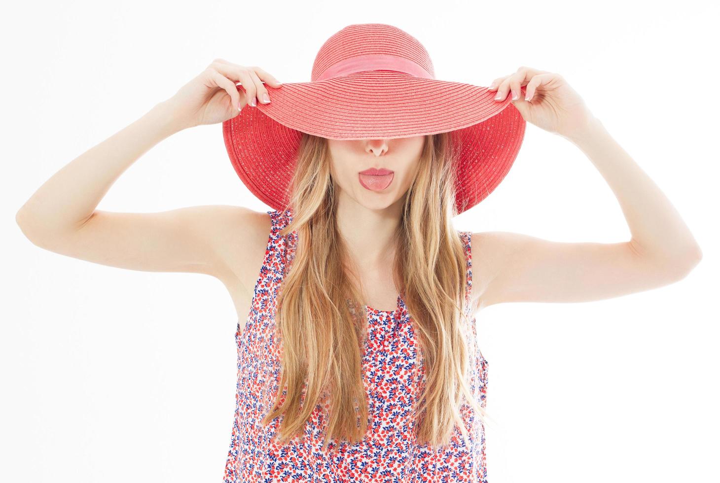 hermosa chica atractiva esconde su rostro detrás de su sombrero de verano y muestra su lengua aislada sobre blanco. foto