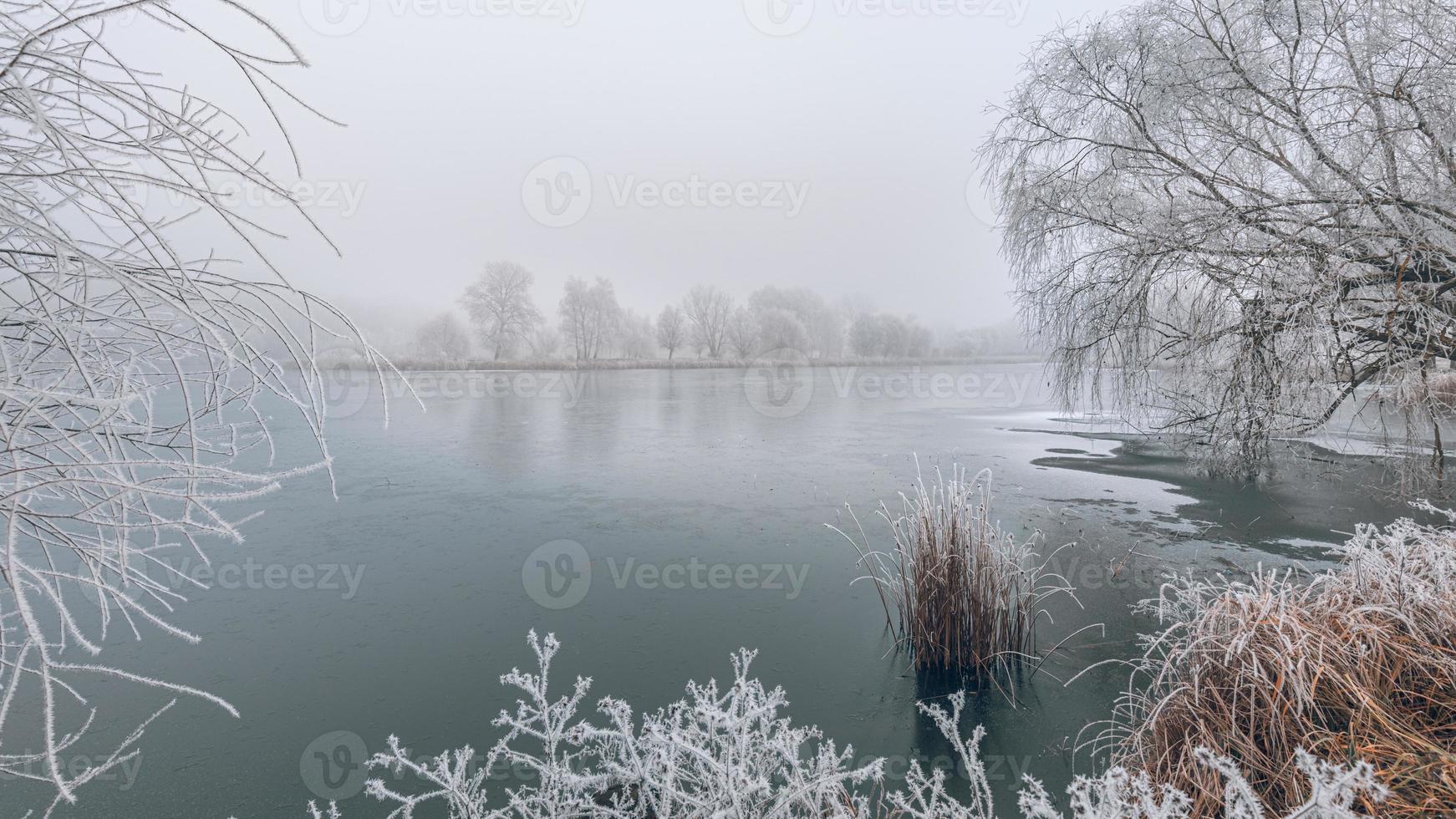 Winter forest and frozen lake sunset. Panoramic landscape with snowy trees, sun, beautiful frozen river with reflection in water. Cold winter landscape artistic foggy morning sunlight. Seasonal nature photo