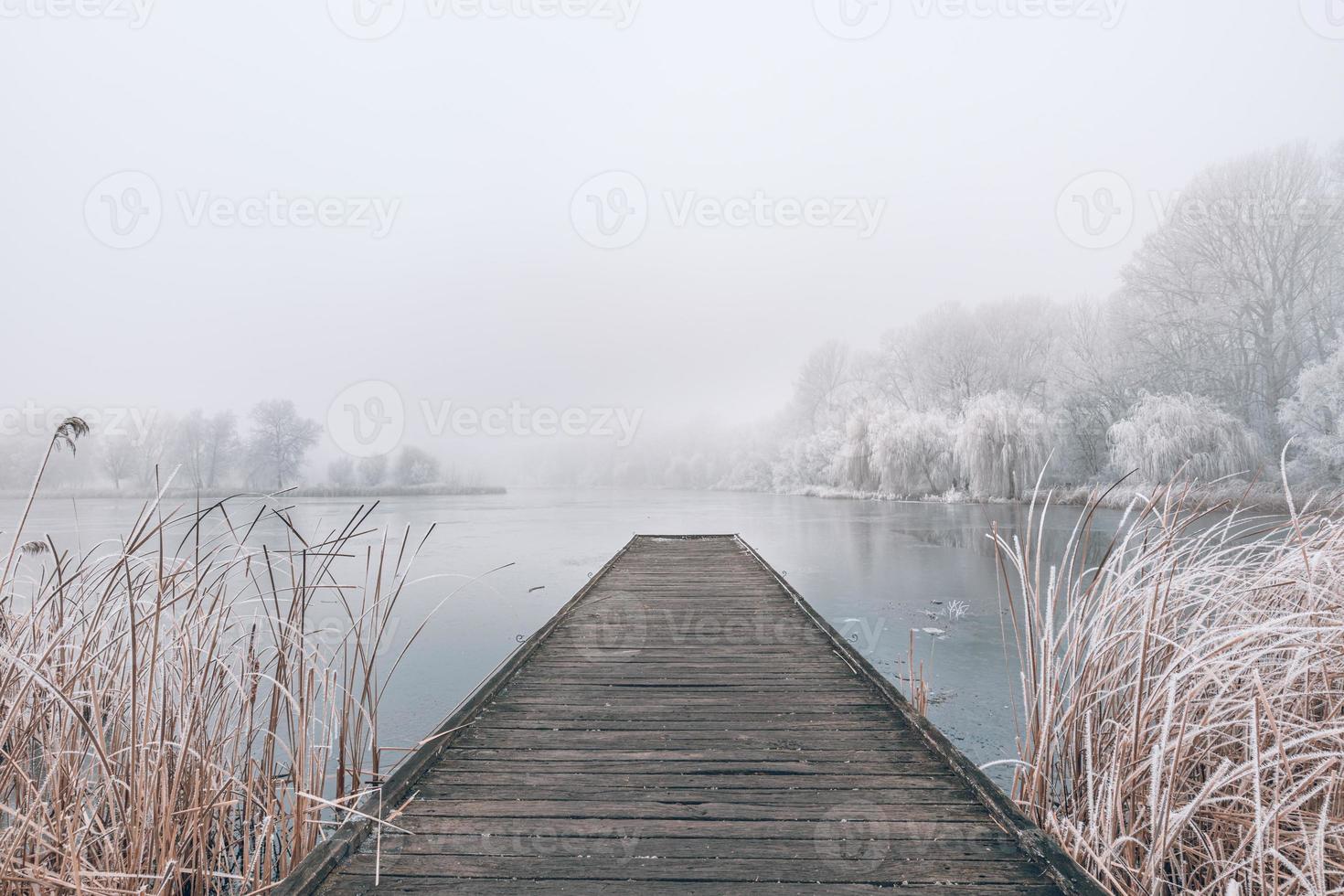 Evening winter landscape. Wooden pier over a beautiful frozen lake. Trees with frost, calm seasonal winter scenery. Peaceful, white view photo