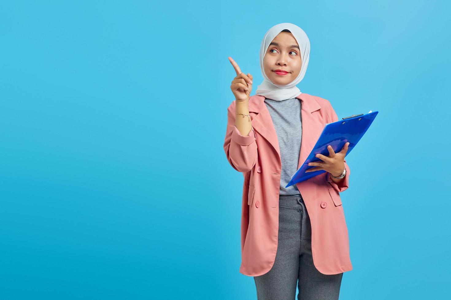 Portrait of a beautiful smiling woman holding a folder standing and pointing to an empty space on a blue background photo