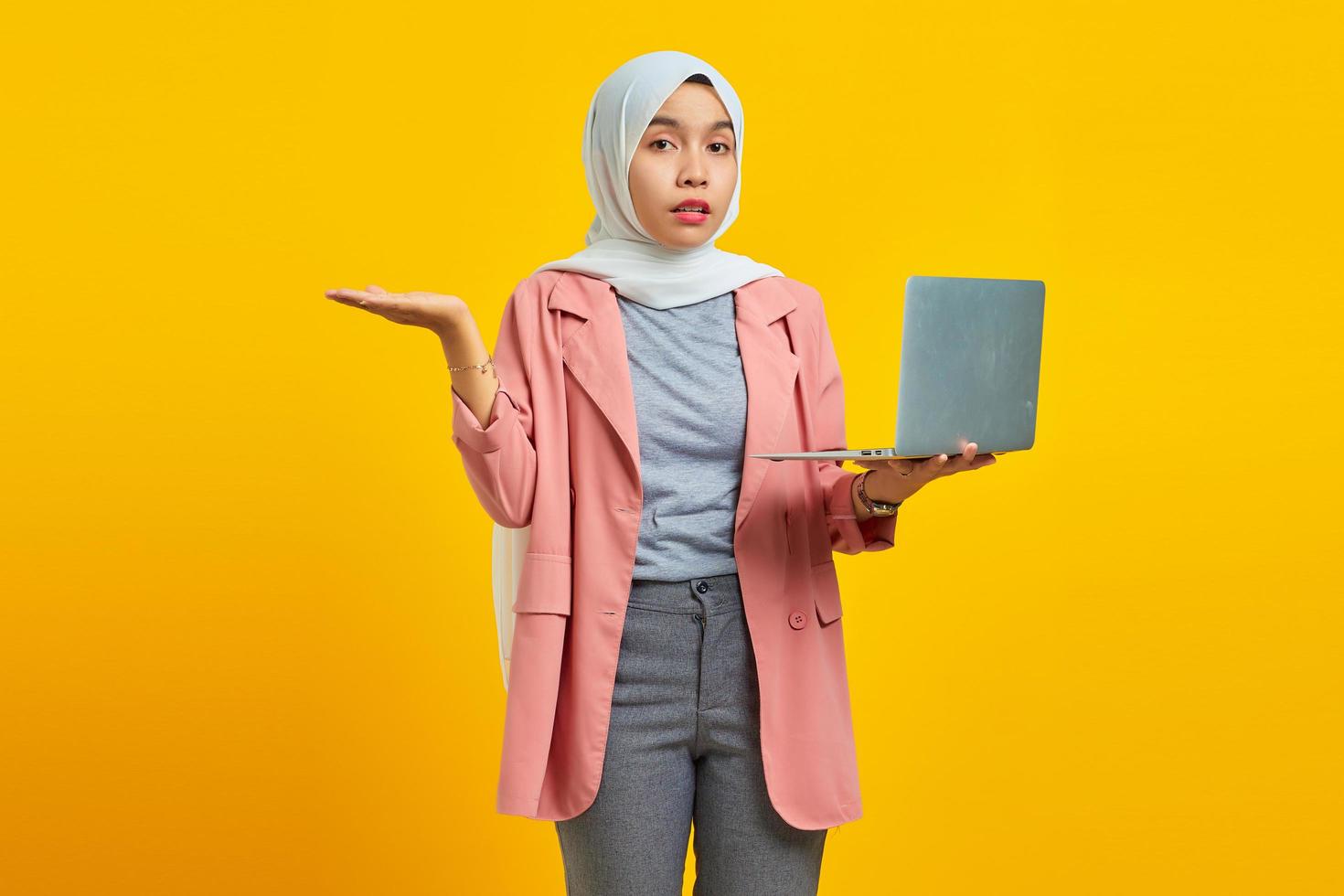 Portrait of asian woman holding laptop and confused isolated over yellow background photo