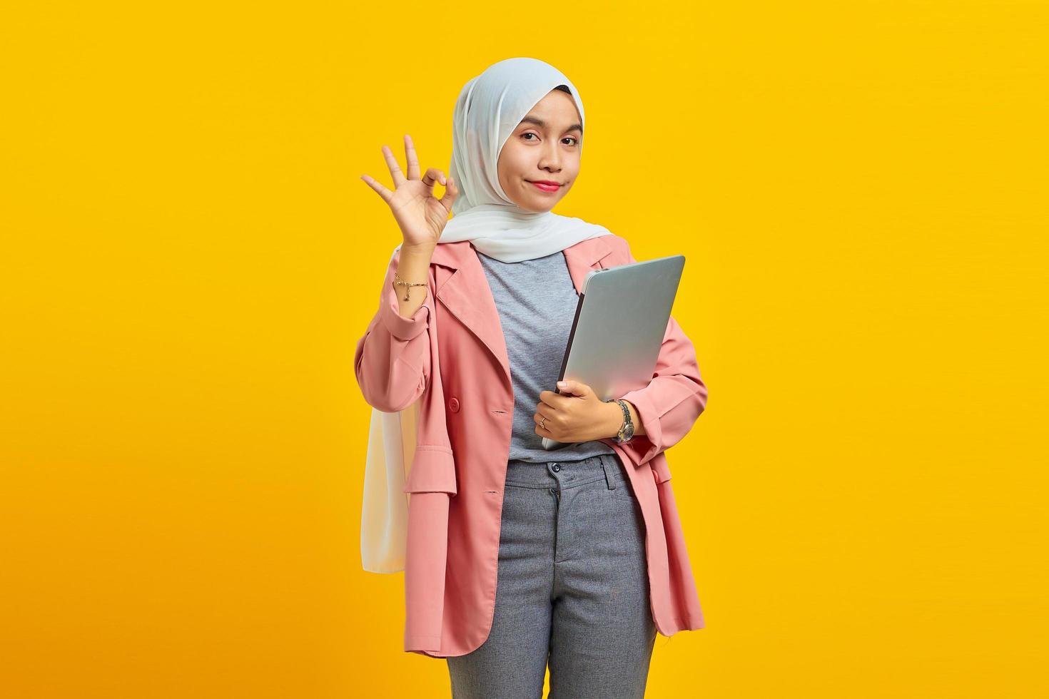 Portrait of a cheerful asian woman holding laptop and making ok gesture isolated over yellow background photo