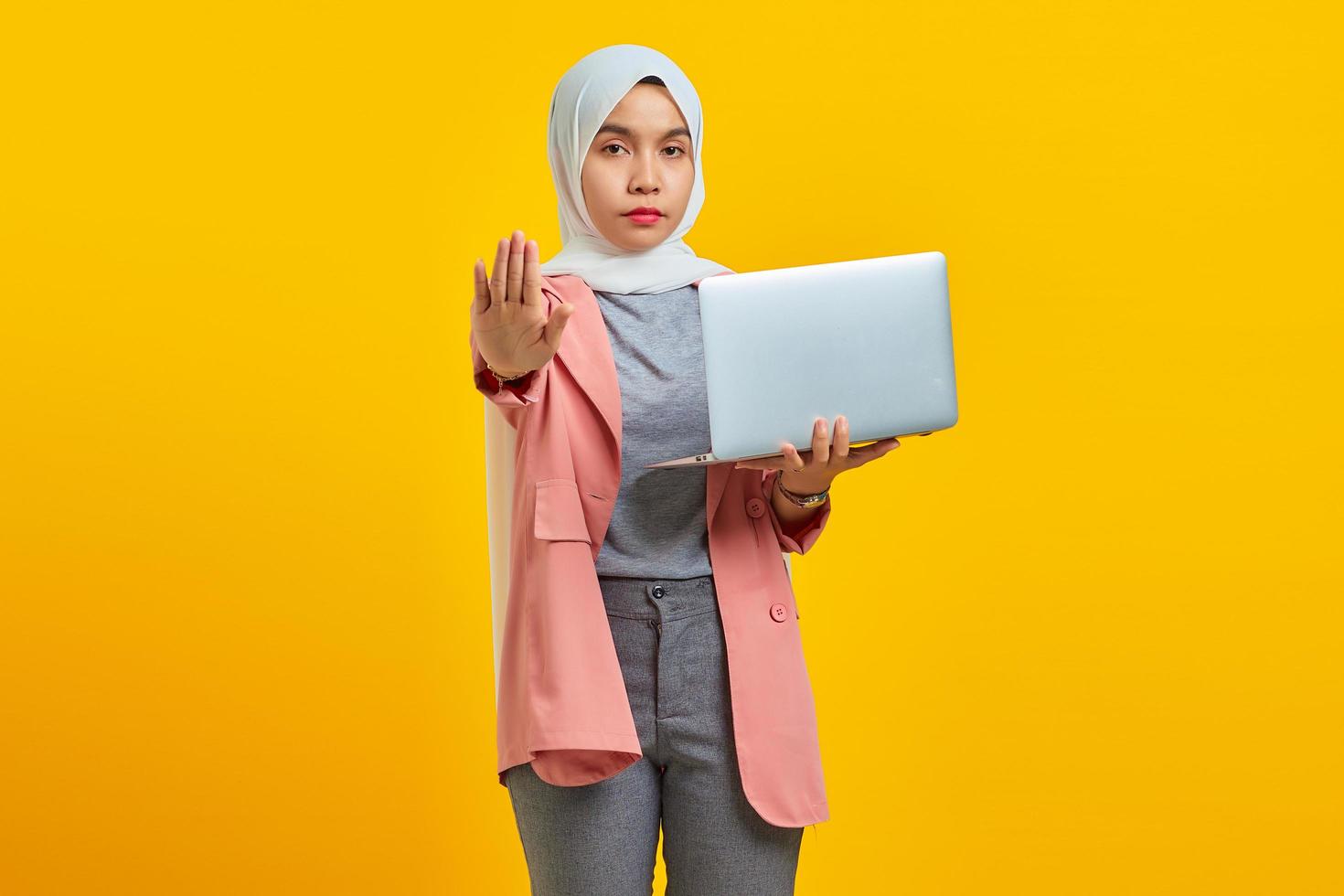 Portrait of beautiful Asian woman holding laptop with open arms doing stop sign with serious and confident expression photo