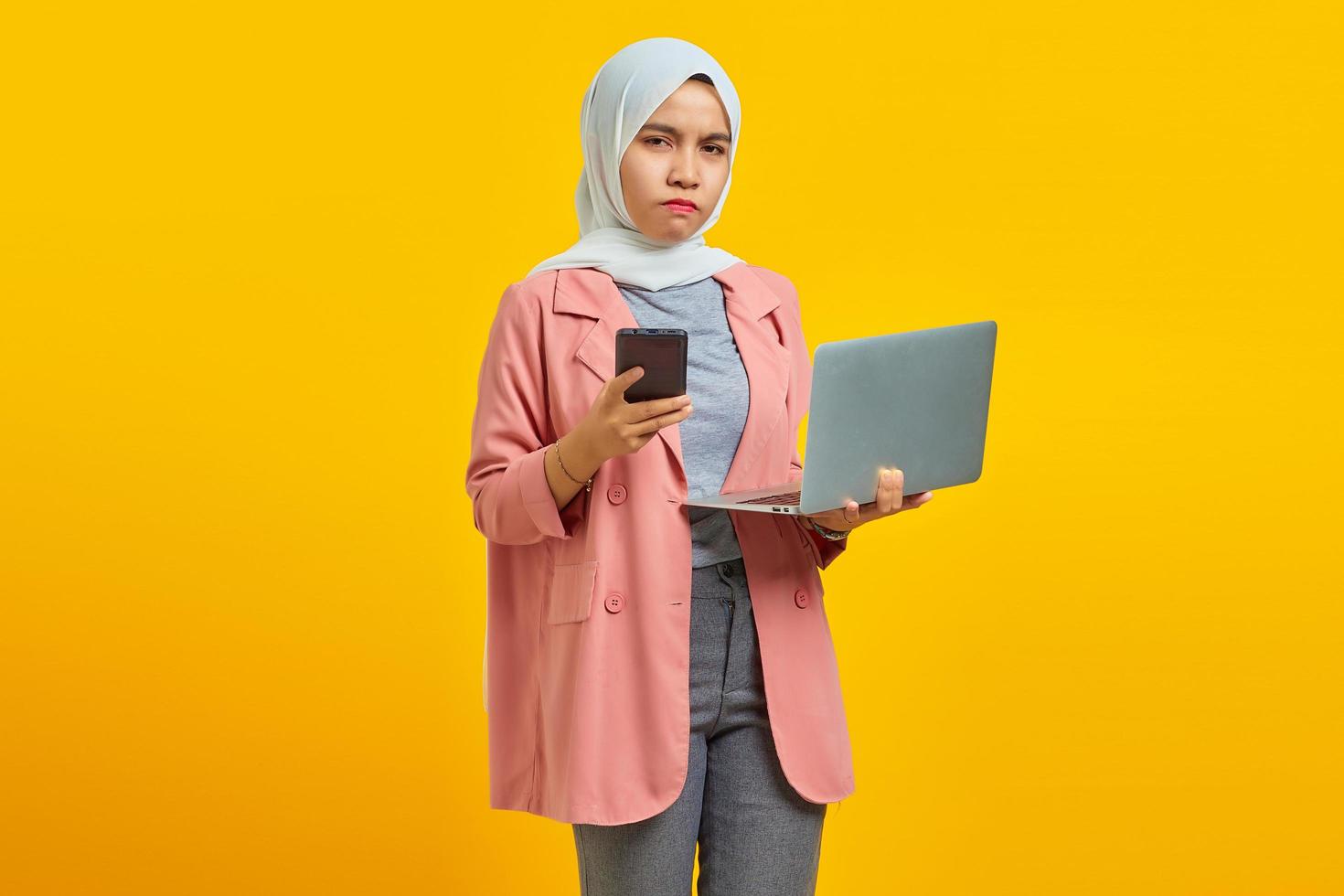 Angry and unhappy young Asian woman holding mobile phone and laptop on blue background photo