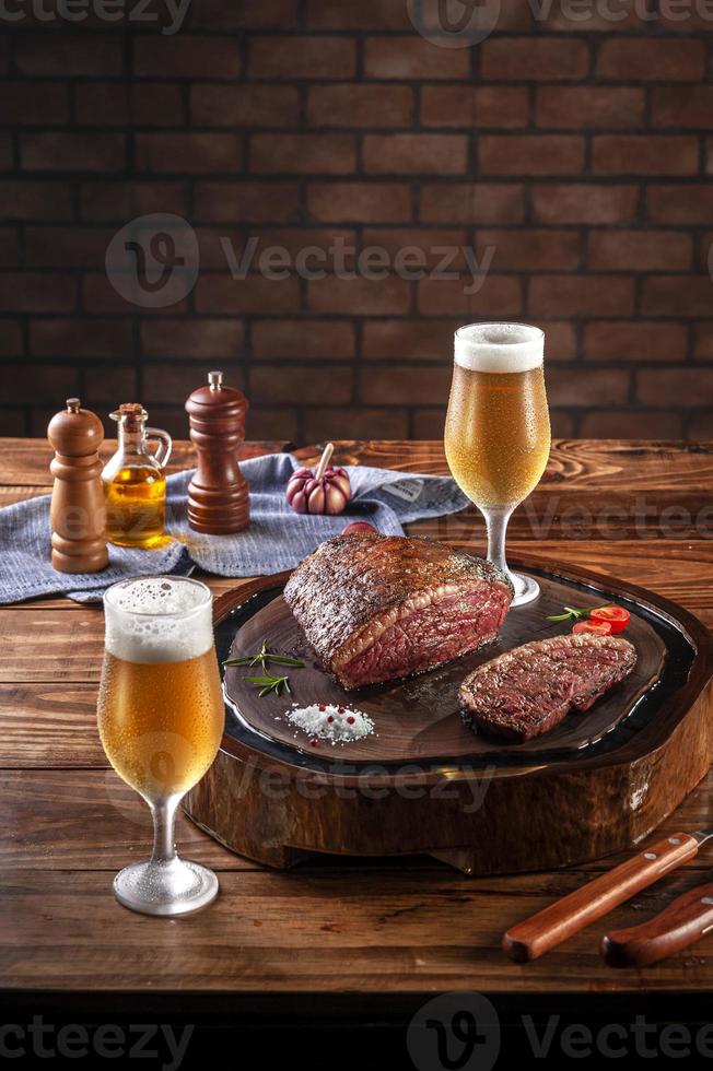 Bistec de grupa a la parrilla sobre tabla de cortar de madera con dos vasos de cerveza tulipa fría sudorosa. mesa de madera y fondo de pared de ladrillos - picanha brasileña. foto