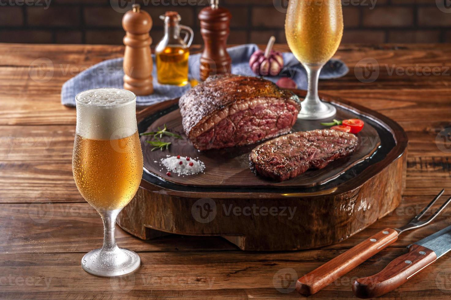 Dos vasos de cerveza tulipa fría sudorosa con bistec de lomo de tapa en rodajas a la parrilla sobre tabla de cortar de madera - picanha brasileña. foto