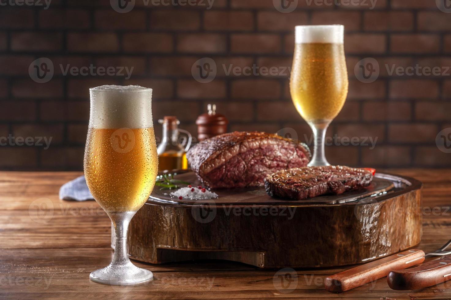 Dos vasos de cerveza tulipa fría sudorosa con bistec de lomo de tapa en rodajas a la parrilla sobre tabla de cortar de madera - picanha brasileña. foto