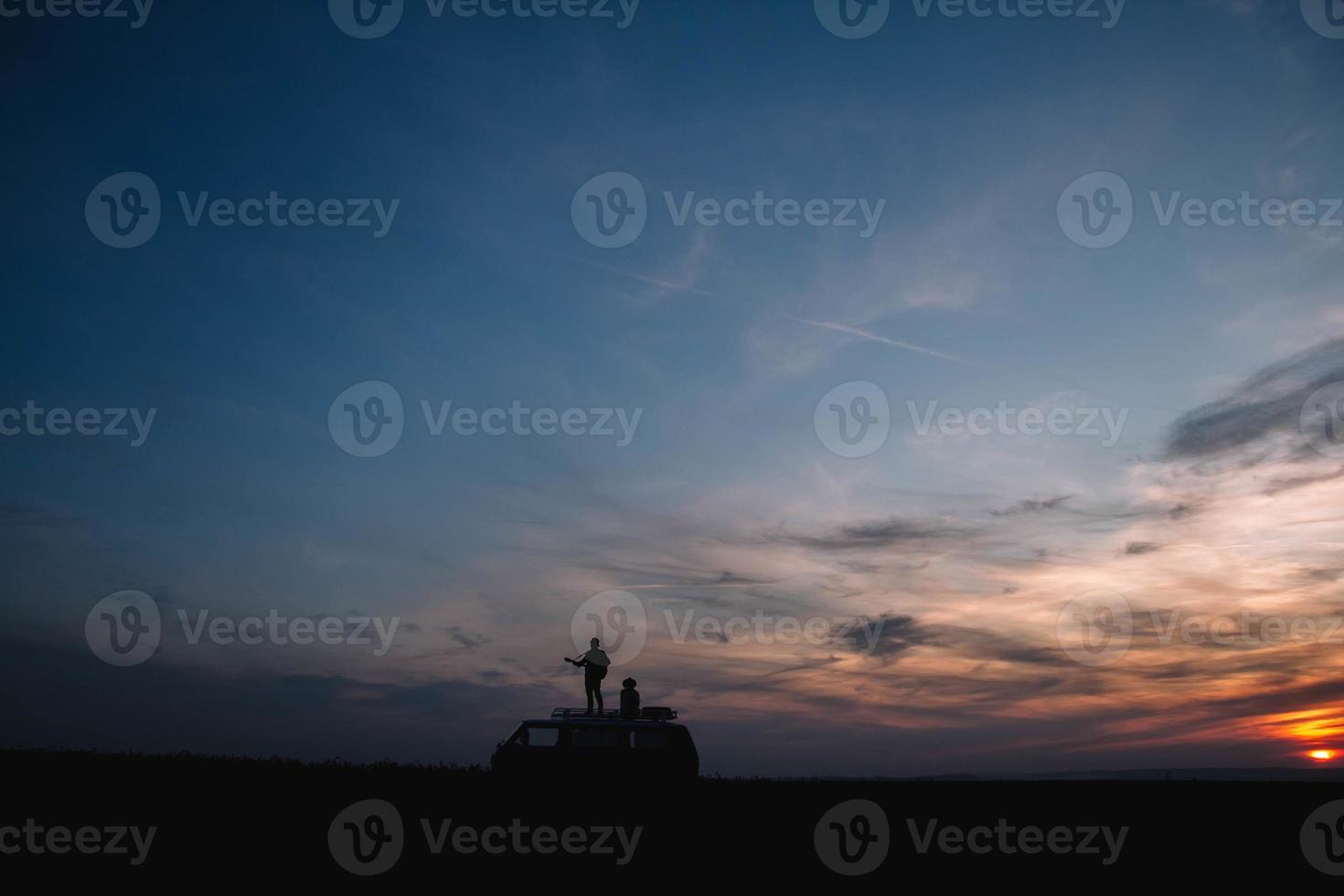 Silhouette of a man with a guitar and a woman in a hat stand on the roof of a car on the background of the sunset photo