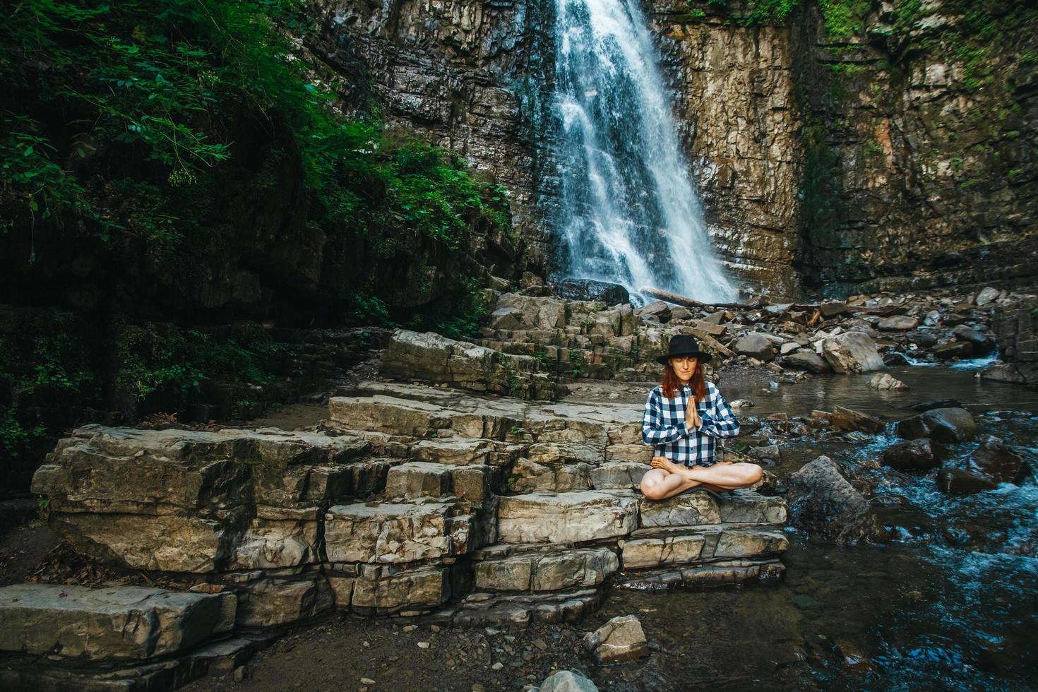 Mujer con cabello rojo con sombrero y camisa meditando sobre rocas en posición de loto contra una cascada foto