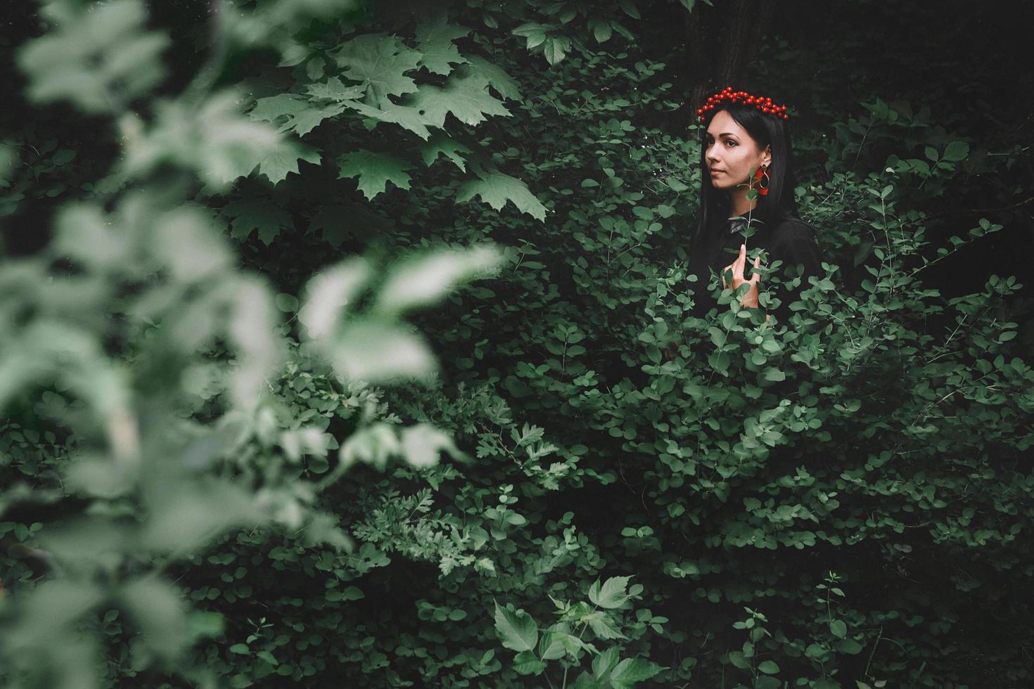 Woman in black dress and red decorations that holds a twig in her hand against background of forest photo