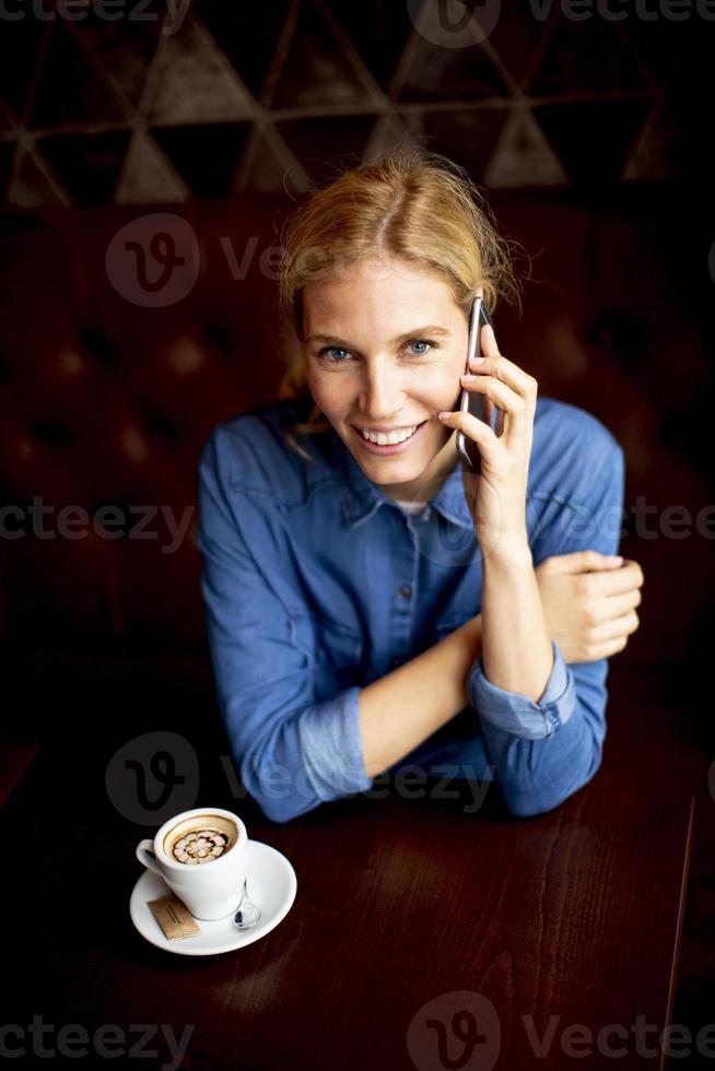 Bastante joven sentada en una mesa con café o capuchino y utilizando un teléfono móvil foto