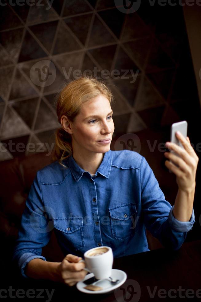 Bastante joven sentada en una mesa con café o capuchino y utilizando un teléfono móvil foto