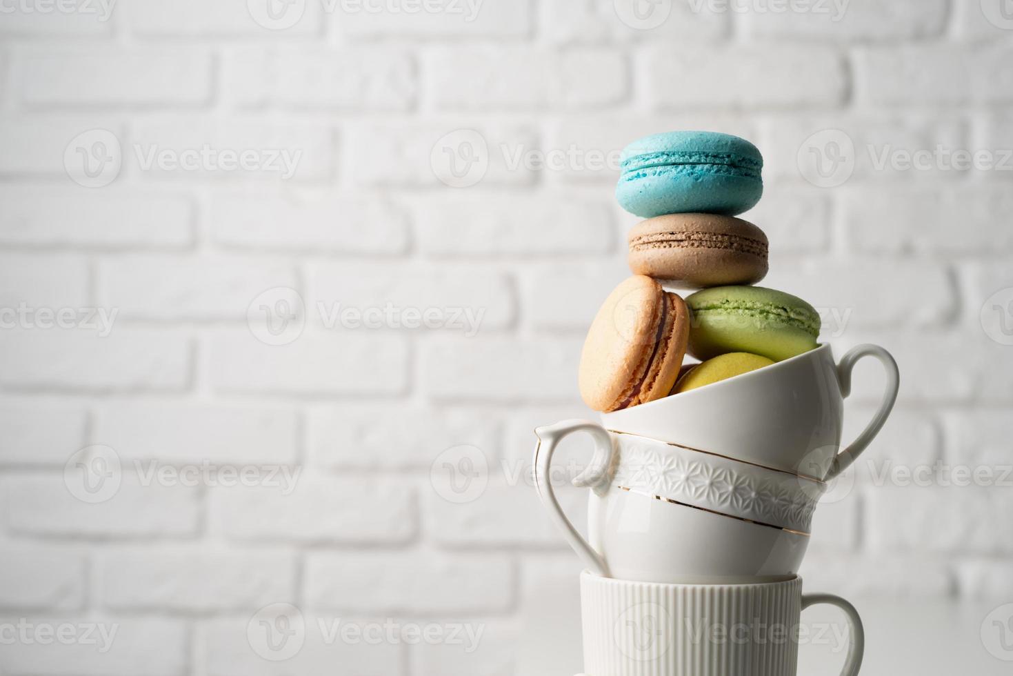 Pila de tazas de café con leche llenas de macarrones en el borde de la mesa, fondo de pared de ladrillo blanco foto